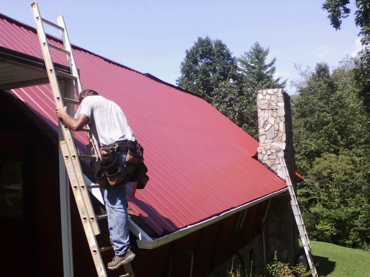 Roofer on a ladder beside a red metal roof and stone chimney — Soffit & Fascia Repair by Roofing Brothers in Jackson Center